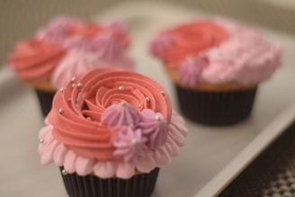 Three cupcakes with intricate pink and purple frosting, decorated with silver edible beads, are displayed on a white tray. The frosting features a rose-like swirl with petal-like details.