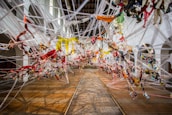 An intricate art installation made of multicolored fabric strips intertwined and suspended in a spacious room, creating a dynamic and chaotic pattern. The installation stretches across the wooden floor, with white arches and walls in the background, illuminated by natural light from high windows.