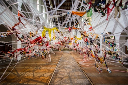 An intricate art installation made of multicolored fabric strips intertwined and suspended in a spacious room, creating a dynamic and chaotic pattern. The installation stretches across the wooden floor, with white arches and walls in the background, illuminated by natural light from high windows.