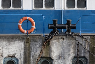 A section of a boat's side featuring a bright orange life preserver attached to a blue railing. Below, a black bollard is secured with heavy chains against a weathered concrete surface. The windowed cabin area above has a clean, blue and white paint job, contrasting with the aging structure beneath.