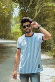 A man wearing sunglasses and a striped t-shirt stands in the middle of a sunlit, tree-lined path. His demeanor is confident, with one hand adjusting his eyewear as he gazes forward.