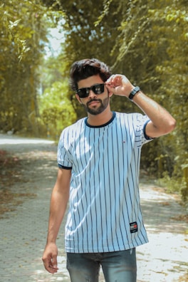 A man wearing sunglasses and a striped t-shirt stands in the middle of a sunlit, tree-lined path. His demeanor is confident, with one hand adjusting his eyewear as he gazes forward.