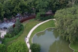 An aerial view of a park area featuring a small pond surrounded by lush greenery and a curved walking path. People are gathered near a building with a glass facade, possibly an entrance or a checkpoint. The scene includes both natural elements like trees and manicured landscapes, as well as human activity.