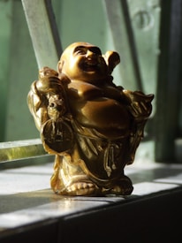 A golden Laughing Buddha figure is placed on a windowsill, with sunlight highlighting its features. The figure is depicted holding a sack over its shoulder and has a joyful expression. The background consists of blurred window bars.