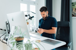 A man standing in a modern office setting is focused on a smartphone while standing at a white desk. The desk is equipped with a large computer monitor, a laptop, and a microphone mounted on an adjustable arm. Potted plants add a touch of greenery to the workspace, and there is a window in the background that lets in natural light.