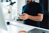 A person is standing at a desk holding a smartphone. There is a microphone hanging from above, a white keyboard, a mouse, and a monitor present. The setting appears to be a modern home office with minimalistic decor and large windows that let in natural light.