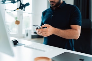 A person is standing at a desk holding a smartphone. There is a microphone hanging from above, a white keyboard, a mouse, and a monitor present. The setting appears to be a modern home office with minimalistic decor and large windows that let in natural light.
