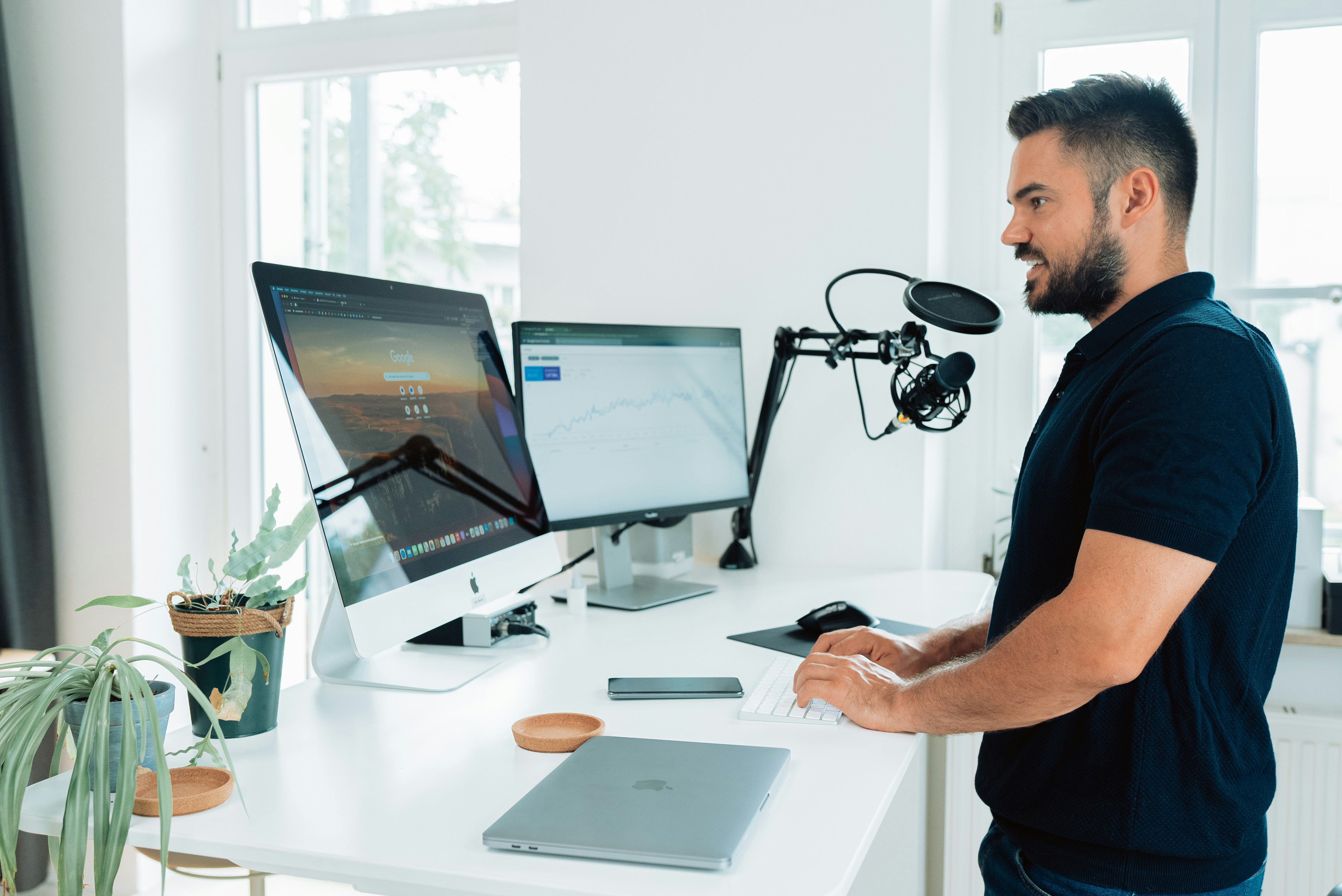 Man at work with podcast microphone in front of a desk and two screens with splitscreen computer and laptop on the desk in a social media agency. Working on a computer.