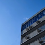 A modern building facade with large blue-tinted glass windows and several air conditioning units mounted on each floor. The sky is clear and vividly blue, dominating the background.