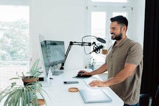 A man standing at a modern, bright home office setup with a desktop computer, microphone on an adjustable arm, and several plants. He is smiling and using a keyboard and mouse, suggesting an engaging, productive environment. The room is well-lit with natural light from windows in the background.