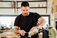 A man with a beard is in a kitchen, pouring ingredients from a jar into a pot on the stove. The kitchen is modern with wooden cabinets, utensils, and various kitchen appliances visible in the background. The setting is bright with natural light coming in through the window.