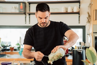 A man with a beard is in a kitchen, pouring ingredients from a jar into a pot on the stove. The kitchen is modern with wooden cabinets, utensils, and various kitchen appliances visible in the background. The setting is bright with natural light coming in through the window.