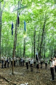 A group of people, possibly scouts, are gathered in a circle in a forest under tall trees. They are wearing uniforms and appear to be involved in some sort of ceremony or activity, with several flags, including a blue and yellow one, hoisted on wooden poles.