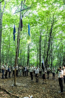 A group of people, possibly scouts, are gathered in a circle in a forest under tall trees. They are wearing uniforms and appear to be involved in some sort of ceremony or activity, with several flags, including a blue and yellow one, hoisted on wooden poles.