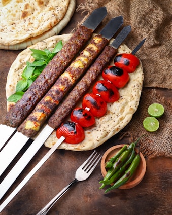A variety of grilled kebabs and roasted tomatoes are presented on skewers resting on flatbread. Fresh basil leaves accompany the kebabs, and fresh lime halves and green chili peppers are placed nearby on a rustic table setting.