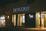 A storefront is illuminated at night, featuring large glass windows and a prominent sign reading 'HEW & DRAW'. The building exterior is modern with a combination of earth-toned bricks and wooden accents. To the left, another shop with a sign 'Picture Frames' is visible, showcasing framed art through its window.