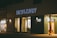 A storefront is illuminated at night, featuring large glass windows and a prominent sign reading 'HEW & DRAW'. The building exterior is modern with a combination of earth-toned bricks and wooden accents. To the left, another shop with a sign 'Picture Frames' is visible, showcasing framed art through its window.