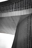 An architectural structure features modern, sleek lines with vertical and horizontal elements. The façade appears to be made of metal panels, with a geometric design that includes curved and flat surfaces. The upper section has rows of windows, and the overall design gives a sense of futuristic and minimalist style.