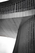 An architectural structure features modern, sleek lines with vertical and horizontal elements. The façade appears to be made of metal panels, with a geometric design that includes curved and flat surfaces. The upper section has rows of windows, and the overall design gives a sense of futuristic and minimalist style.