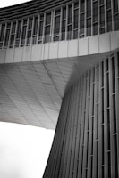 An architectural structure features modern, sleek lines with vertical and horizontal elements. The façade appears to be made of metal panels, with a geometric design that includes curved and flat surfaces. The upper section has rows of windows, and the overall design gives a sense of futuristic and minimalist style.