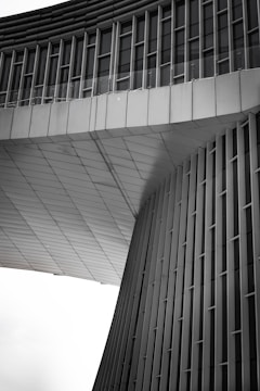An architectural structure features modern, sleek lines with vertical and horizontal elements. The façade appears to be made of metal panels, with a geometric design that includes curved and flat surfaces. The upper section has rows of windows, and the overall design gives a sense of futuristic and minimalist style.