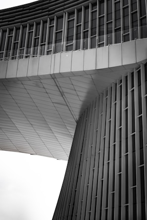 An architectural structure features modern, sleek lines with vertical and horizontal elements. The façade appears to be made of metal panels, with a geometric design that includes curved and flat surfaces. The upper section has rows of windows, and the overall design gives a sense of futuristic and minimalist style.