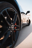 A low-angle shot of a sleek, modern car focusing on its front wheel with a shiny black rim, featuring an emblem at the center. The background is slightly blurred, highlighting the detailed design of the vehicle.