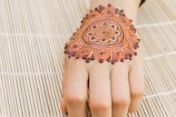 A henna design is intricately drawn on the back of a hand, showcasing detailed patterns with floral and circular motifs. The hand rests on a bamboo mat, providing a textured background.