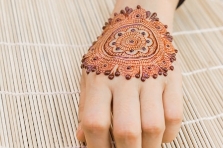 A henna design is intricately drawn on the back of a hand, showcasing detailed patterns with floral and circular motifs. The hand rests on a bamboo mat, providing a textured background.