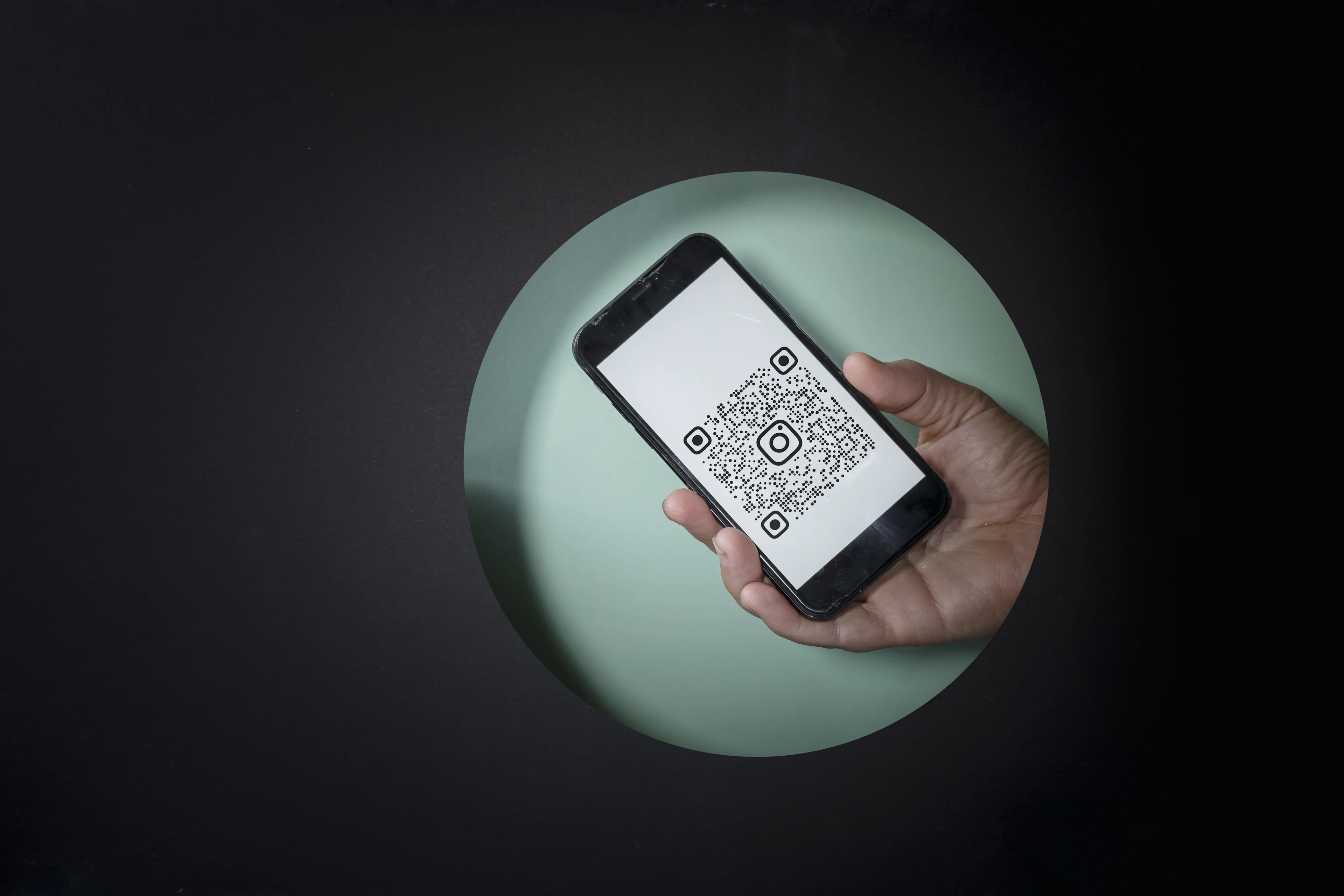 A hand holding a smartphone that shows a black-and-white QR code on its screen, seen through a round cutout in a dark background