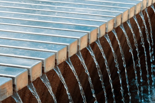 Water flows uniformly over the edge of a structure composed of evenly spaced, parallel metal bars into a basin below.