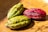 Three cacao pods rest on a surface illuminated by sunlight. The surface appears to be brown and slightly wet. Two of the pods are green with dark patches, while the third pod is red with a similar texture.