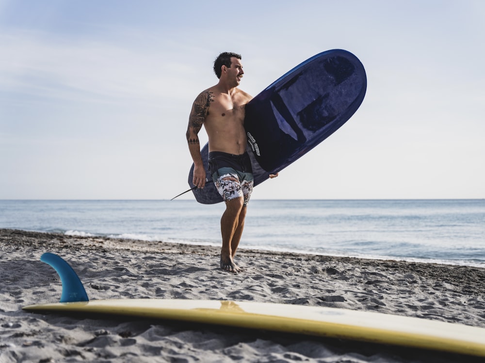 uomo in pantaloncini blu che tiene la tavola da surf blu sulla spiaggia durante il giorno