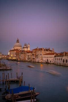A serene evening scene of a canal in Venice, dominated by historic architecture. The calm waters reflect the subtle colors of the sunset, with boats gently moving through the canal. Domed buildings and illuminated windows create a picturesque atmosphere.