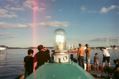 An outdoor scene by the water features a mix of people gathered around an area with a nautical atmosphere. A prominent light fixture in the foreground draws the eye, while the background includes clear skies, scattered clouds, and several boats. The water reflects the light and movement of a peaceful day.
