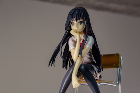 A figurine of an anime character with long black hair and a contemplative expression sits on a chair. The character is wearing a school uniform consisting of a white shirt and dark-colored skirt. The background is neutral, drawing focus to the figurine.