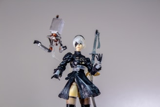 A detailed figure of a character with short white hair and a black outfit, including a dress and thigh-high boots, stands prominently. The figure is accompanied by a small mechanical floating object with articulated limbs. An ornate sword is visible in the background.