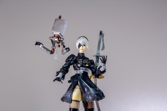 A detailed figure of a character with short white hair and a black outfit, including a dress and thigh-high boots, stands prominently. The figure is accompanied by a small mechanical floating object with articulated limbs. An ornate sword is visible in the background.