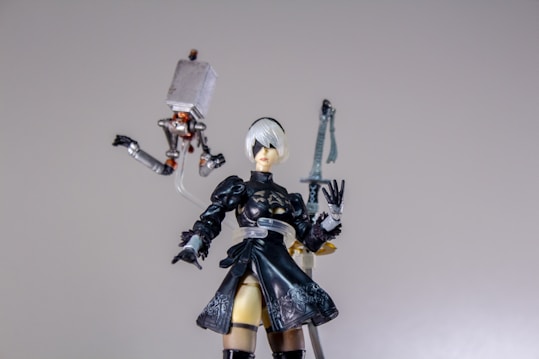 A detailed figure of a character with short white hair and a black outfit, including a dress and thigh-high boots, stands prominently. The figure is accompanied by a small mechanical floating object with articulated limbs. An ornate sword is visible in the background.