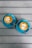 Two cappuccino cups are placed on a wooden table. The cappuccinos have latte art text, with one reading 'COFFEE CHAT?' and the other saying 'YEAH!' Both cups are served in matching blue saucers with silver spoons.