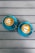Two cappuccino cups are placed on a wooden table. The cappuccinos have latte art text, with one reading 'COFFEE CHAT?' and the other saying 'YEAH!' Both cups are served in matching blue saucers with silver spoons.