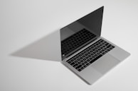 A sleek, open silver laptop with a reflective screen casting a shadow on a plain, light background. The keyboard is visible, with black keys featuring a minimalist design.
