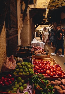 A vibrant market scene with various fresh vegetables and fruits displayed in baskets, such as tomatoes, green peppers, and potatoes. The market is in an alleyway with weathered walls and overhead coverings providing shade. Several people walk through the narrow path, giving the place a bustling, lively atmosphere.