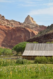 A rustic wooden cabin sits amidst lush green vegetation, with a backdrop of towering, reddish rock formations. The sky is clear with a few wispy clouds, creating a serene and picturesque landscape.
