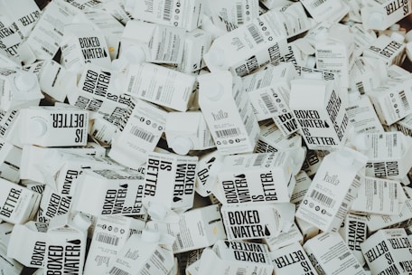 A large number of white cartons with black text saying 'Boxed Water is Better' are piled together. The cartons have graphics and text promoting sustainability.