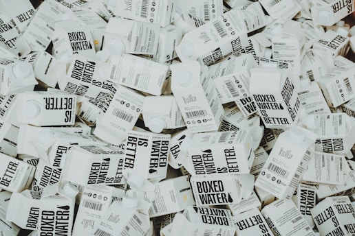 A large number of white cartons with black text saying 'Boxed Water is Better' are piled together. The cartons have graphics and text promoting sustainability.