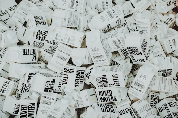 A large number of white cartons with black text saying 'Boxed Water is Better' are piled together. The cartons have graphics and text promoting sustainability.