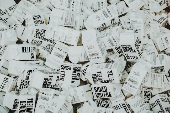 A large number of white cartons with black text saying 'Boxed Water is Better' are piled together. The cartons have graphics and text promoting sustainability.