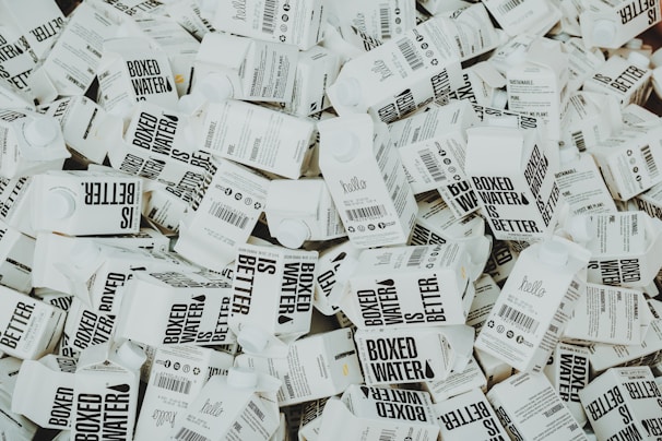 A large number of white cartons with black text saying 'Boxed Water is Better' are piled together. The cartons have graphics and text promoting sustainability.