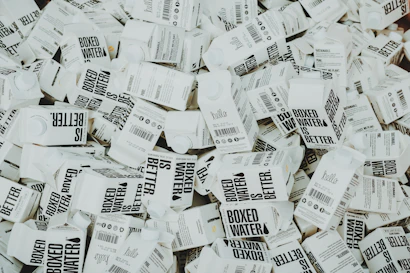 A large number of white cartons with black text saying 'Boxed Water is Better' are piled together. The cartons have graphics and text promoting sustainability.